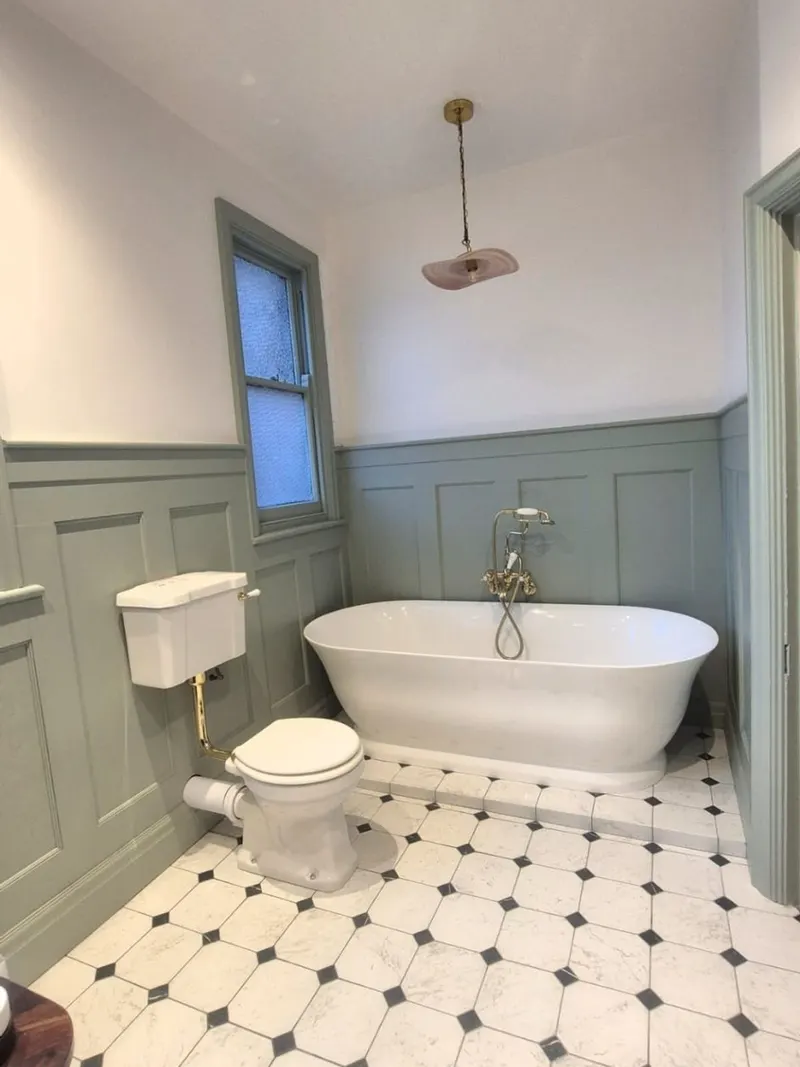 A freestanding bath against sage green panelling with a brass bath shower mixer and octagonal floor tiling
