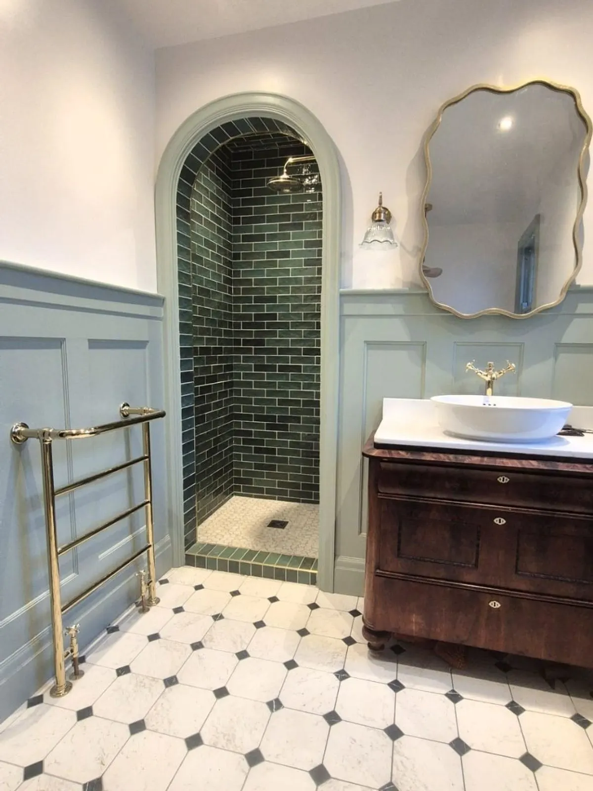 An arched shower recess tiled in dark green metro tile beside a vanity unit and brass heated towel rail