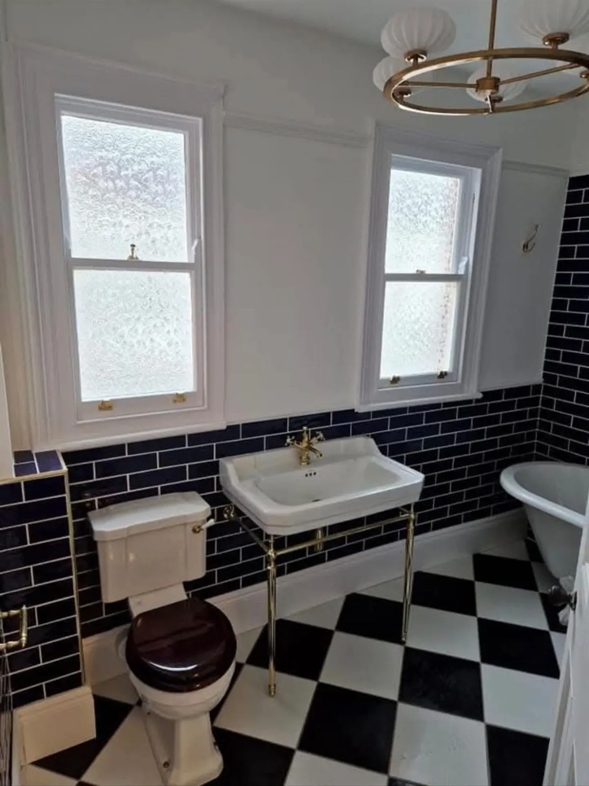 A basin on brass legs beneath two sash windows, with navy tiling and a brass ceiling light