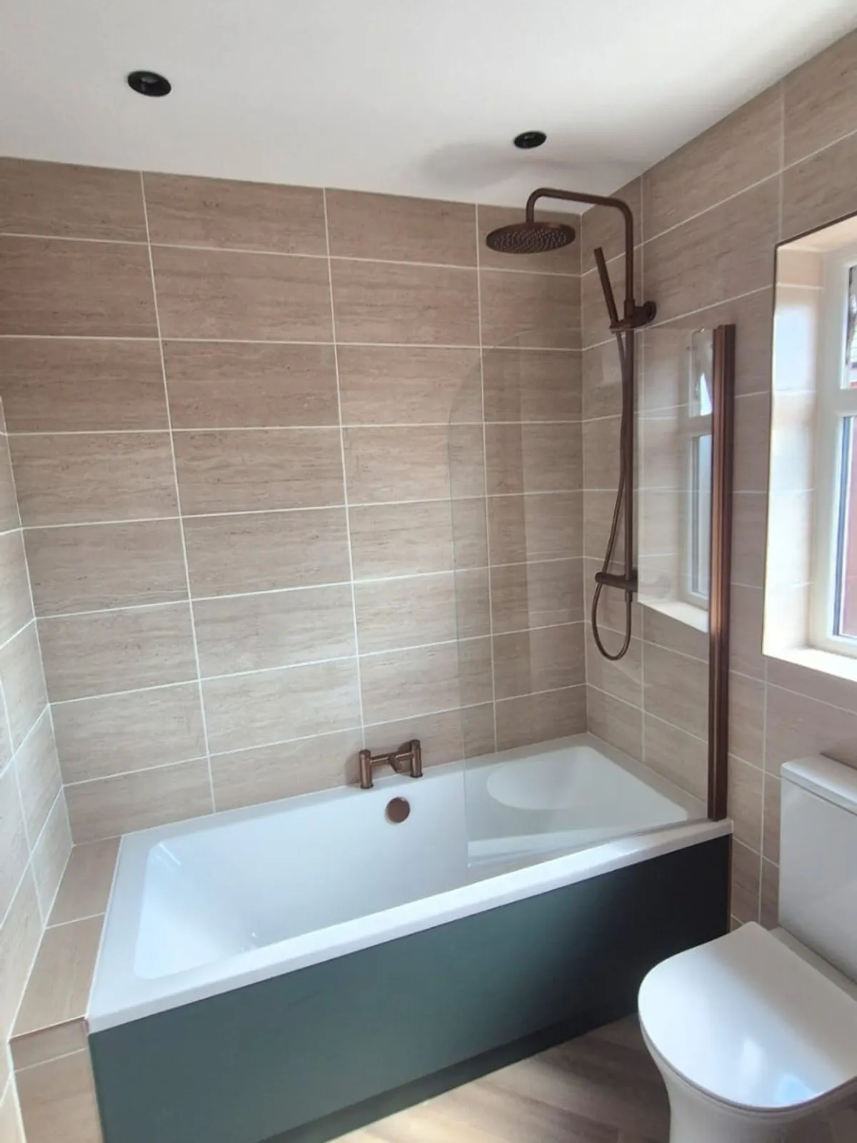 A refitted bathroom with wood effect wall tile, a green bath panel and a copper rainfall shower